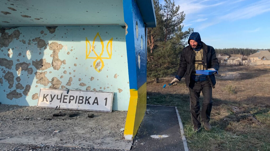 Person painting a bus stop with blue and yellow colors in a rural area. The structure has visible wear, and a sign reading "Кучерівка 1" lies on the ground.