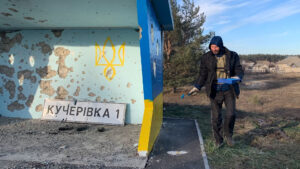 Person painting a bus stop with blue and yellow colors in a rural area. The structure has visible wear, and a sign reading "Кучерівка 1" lies on the ground.