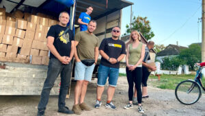A group of people stands in front of an open truck loaded with boxes. Two individuals are inside the truck, while others are standing on the ground.