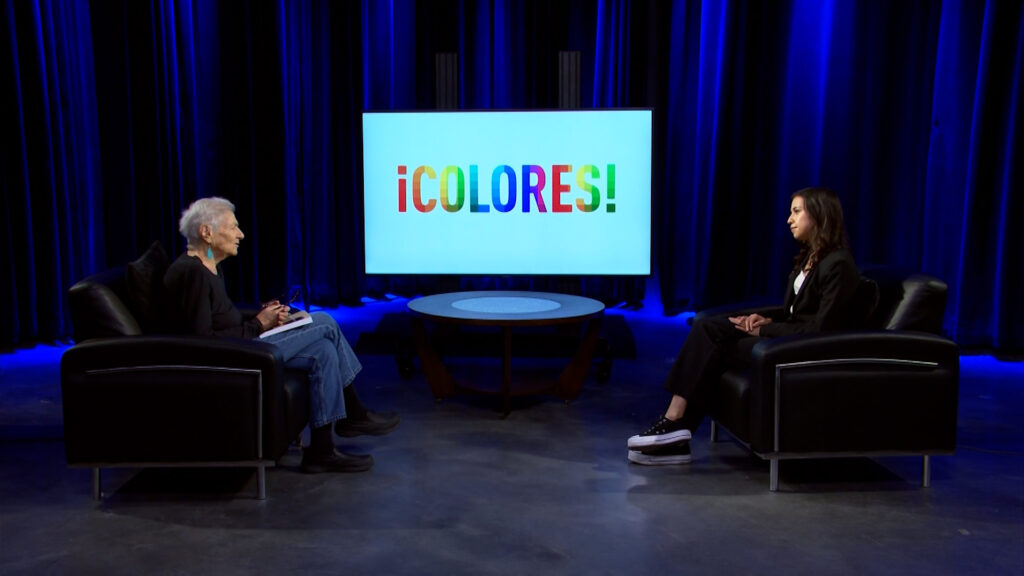 Two people seated in black armchairs facing each other in a studio with a blue backdrop. A screen between them displays the word "¡COLORES!" in colorful letters.