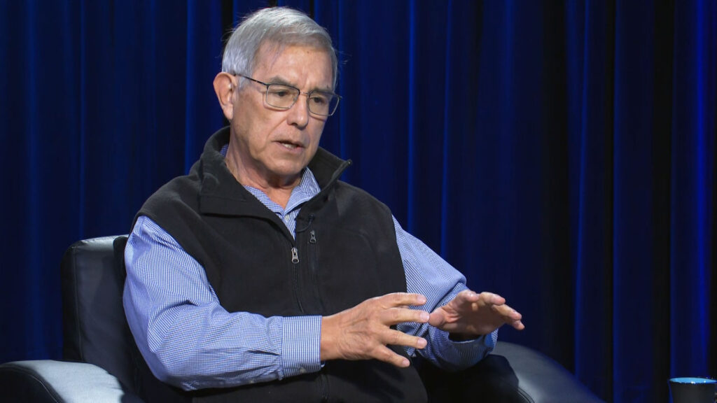 An older man wearing glasses, a black vest, and a blue shirt gestures with his hands while sitting in a black chair against a dark blue curtain background.