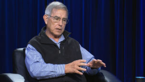An older man wearing glasses, a black vest, and a blue shirt gestures with his hands while sitting in a black chair against a dark blue curtain background.