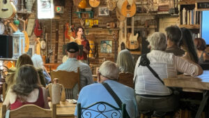 A woman stands at a microphone reading to an audience seated in a cozy, guitar-filled room with eclectic decorations and wooden furniture.