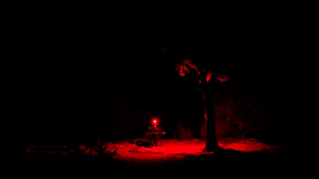 A dimly lit outdoor scene at night shows a person sitting by a table under a tree, illuminated by a red light.