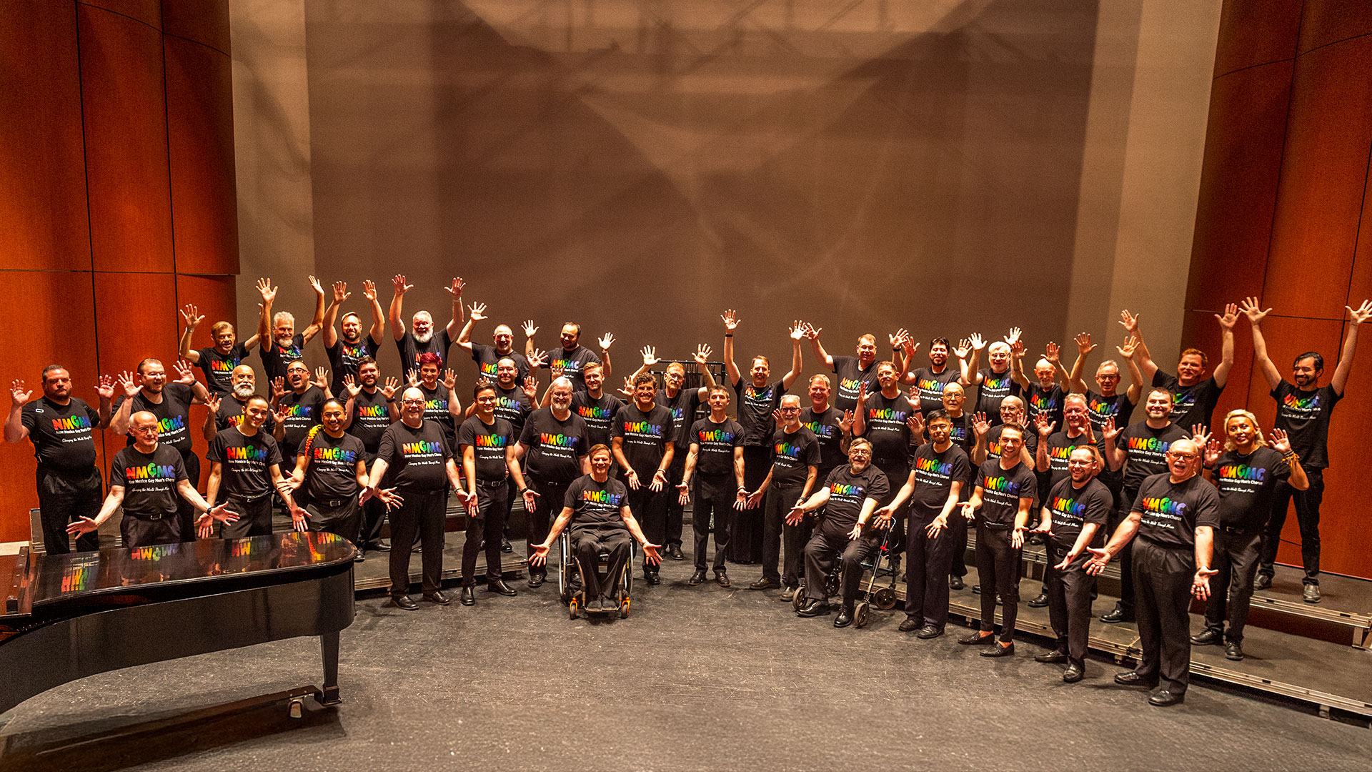 A large choir of diverse adults, some in wheelchairs, pose on stage wearing matching black shirts with rainbow lettering, arms raised, in front of a grand piano and wood-paneled background.