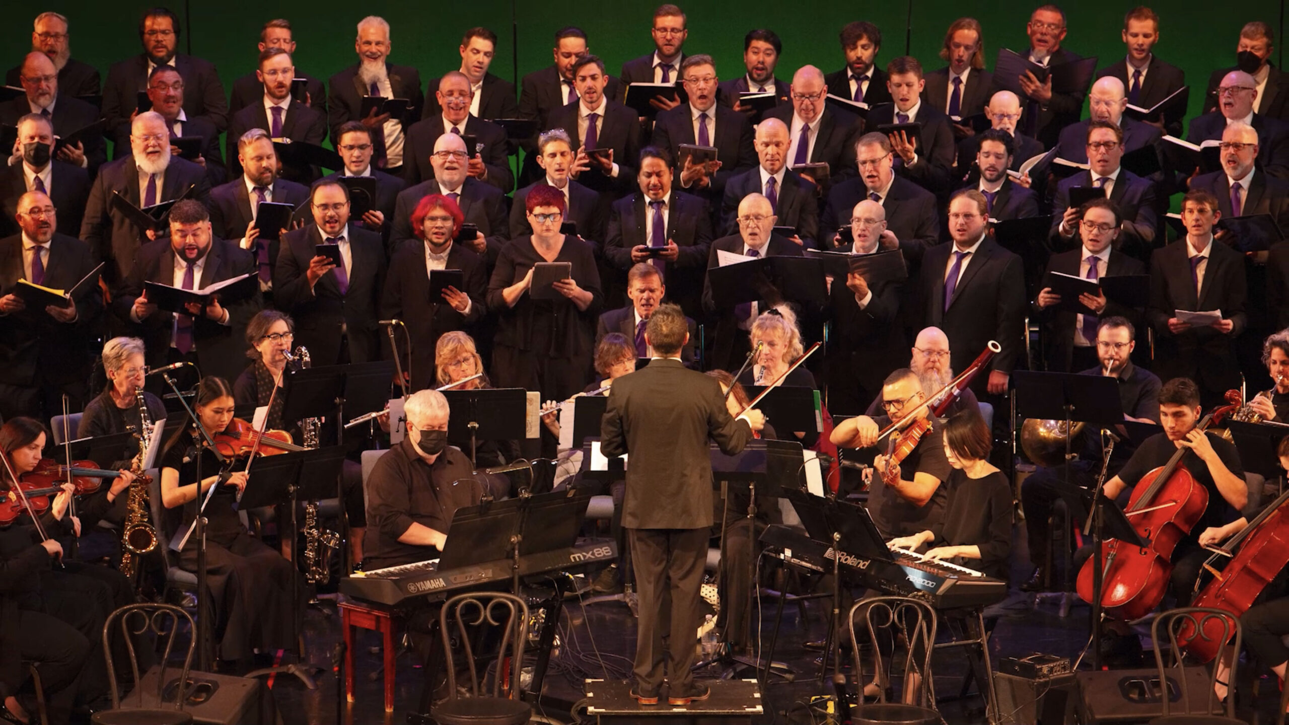 A conductor leads an orchestra and choir in performance; musicians are seated with instruments and sheet music, while choristers stand behind them singing.