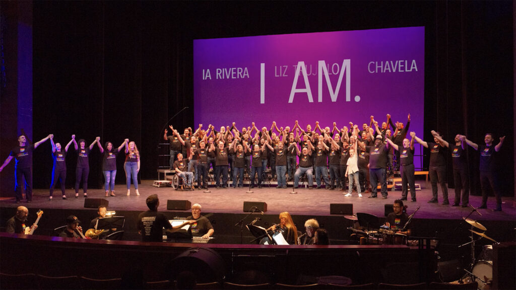 A large choir stands on stage with raised hands, accompanied by a seated orchestra below. The backdrop displays "I AM." and several names in capital letters.