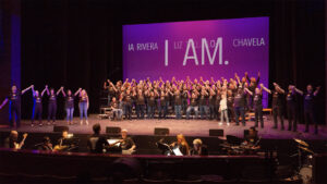 A large choir stands on stage with raised hands, accompanied by a seated orchestra below. The backdrop displays "I AM." and several names in capital letters.