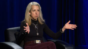 A woman with long blonde hair sits in a black chair, wearing a black turtleneck and maroon pants, gesturing with both hands in front of a dark blue curtain.