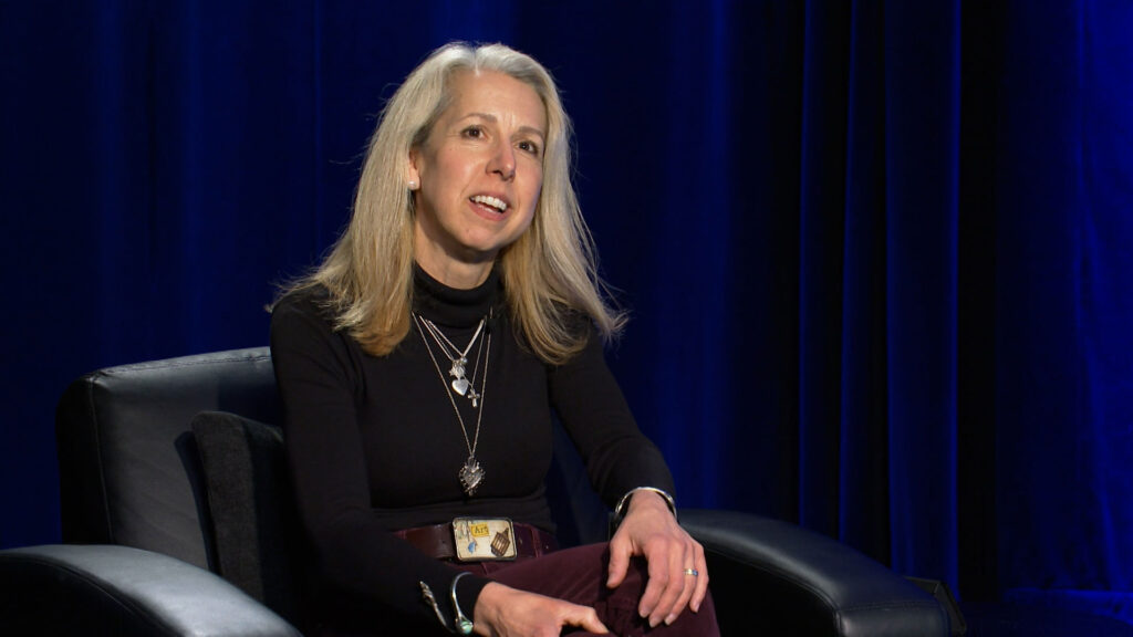 A woman with long blonde hair sits in a black chair against a dark blue curtain, wearing a black turtleneck and several necklaces.