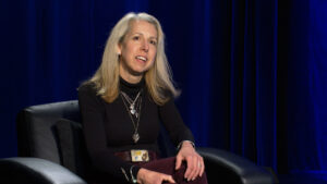 A woman with long blonde hair sits in a black chair against a dark blue curtain, wearing a black turtleneck and several necklaces.