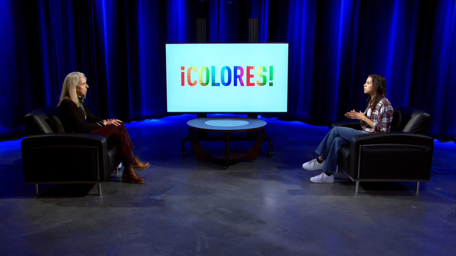 Two women sit across from each other in a studio with blue lighting, a round table between them, and a large screen behind displaying the word "¡COLORES!" in colorful letters.