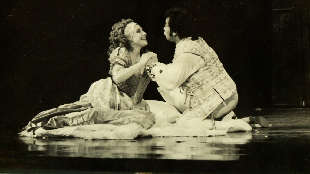 Two actors in period costumes kneel on stage, holding hands and looking at each other, in a theatrical performance.