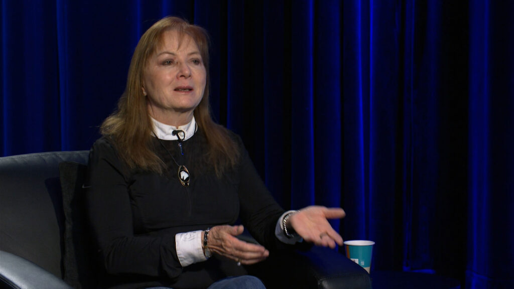 A woman with long, light brown hair sits in a black chair, gesturing with her hands, with a blue curtain and a paper cup visible in the background.