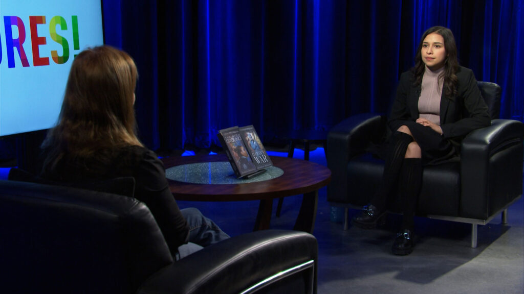 Two women sit in black armchairs facing each other in a studio with blue lighting, a round table and framed photos between them, and a colorful sign partially visible on the left.