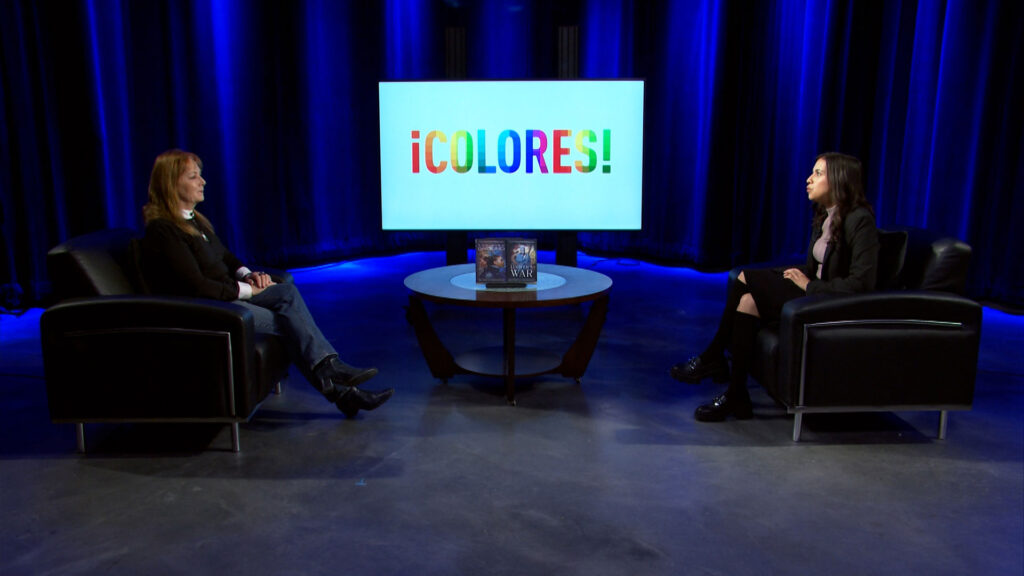 Two women sit across from each other in black chairs on a studio set with a round table and a screen displaying the word "¡COLORES!" in colorful letters.