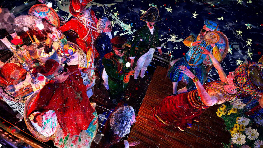 A group of people in colorful, detailed costumes and hats are gathered at a festive event, some are seated at a decorated table while others stand or dance nearby on a wooden floor.