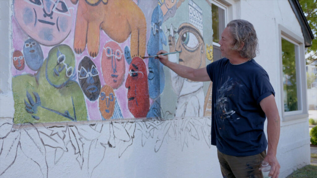 A man paints a colorful mural with various abstract faces and animals on an outdoor white wall, holding a paintbrush and a paint container.