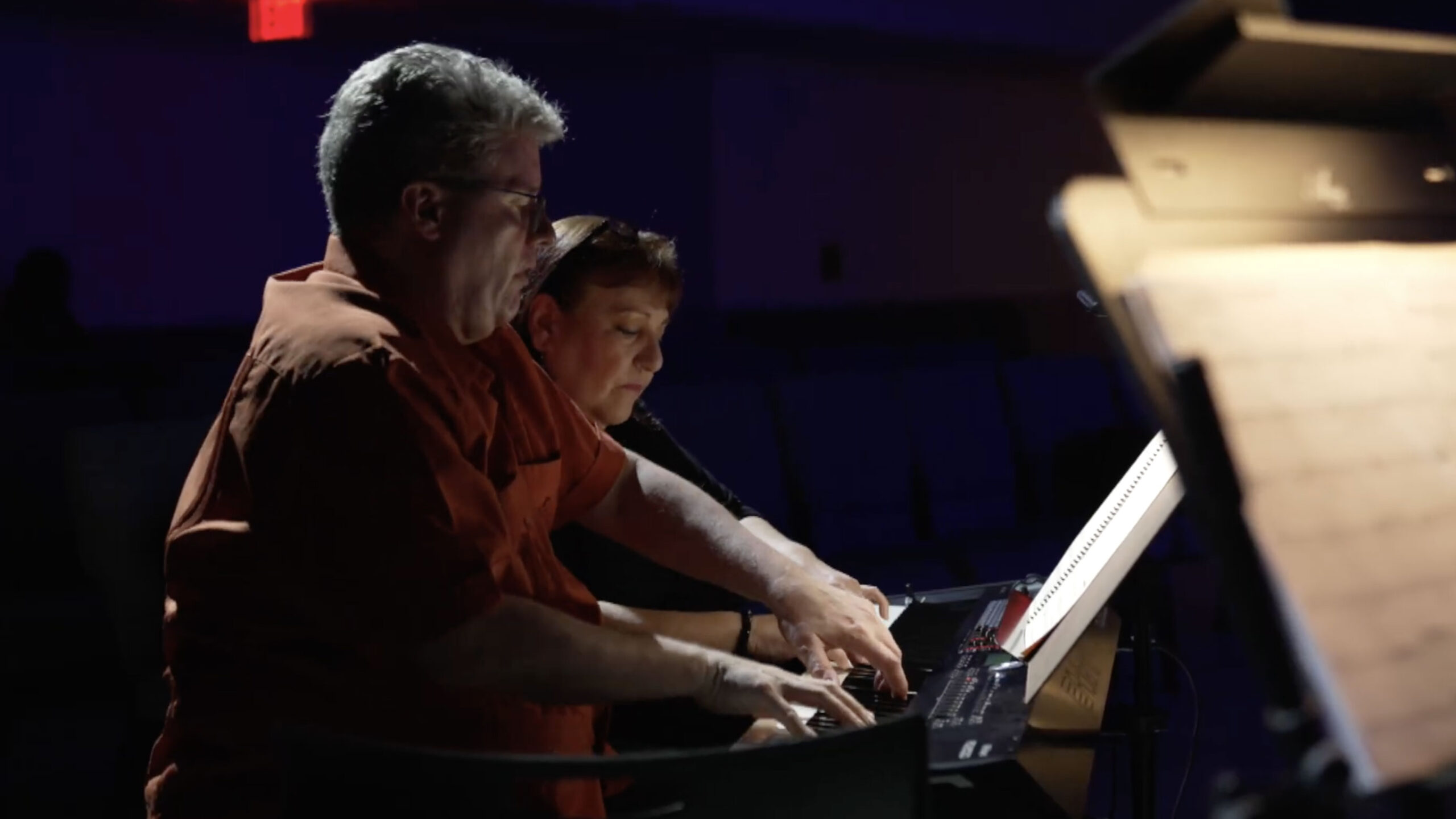 Two people sit side by side playing electronic keyboards, focused on sheet music in a dimly lit room.
