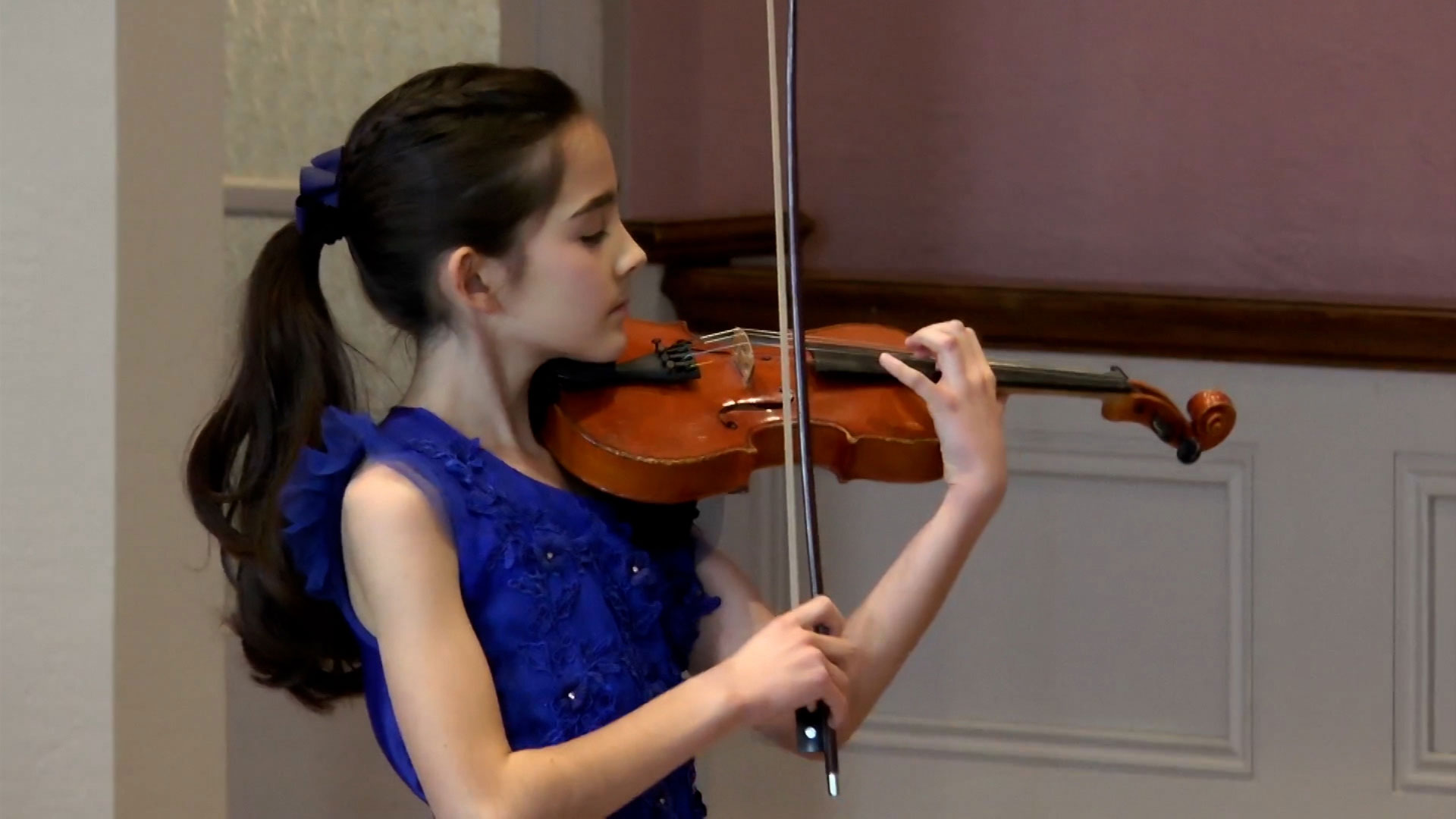 A girl in a blue dress plays the violin indoors, focusing on her performance.