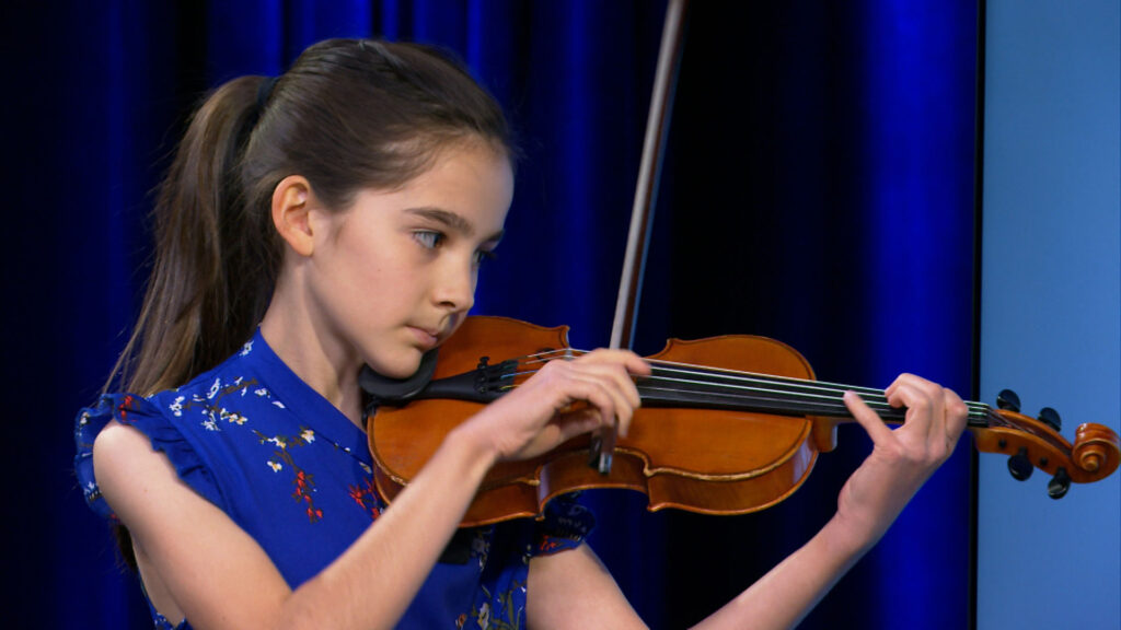 A young girl with long brown hair in a ponytail plays a violin, wearing a blue floral dress, against a blue background.