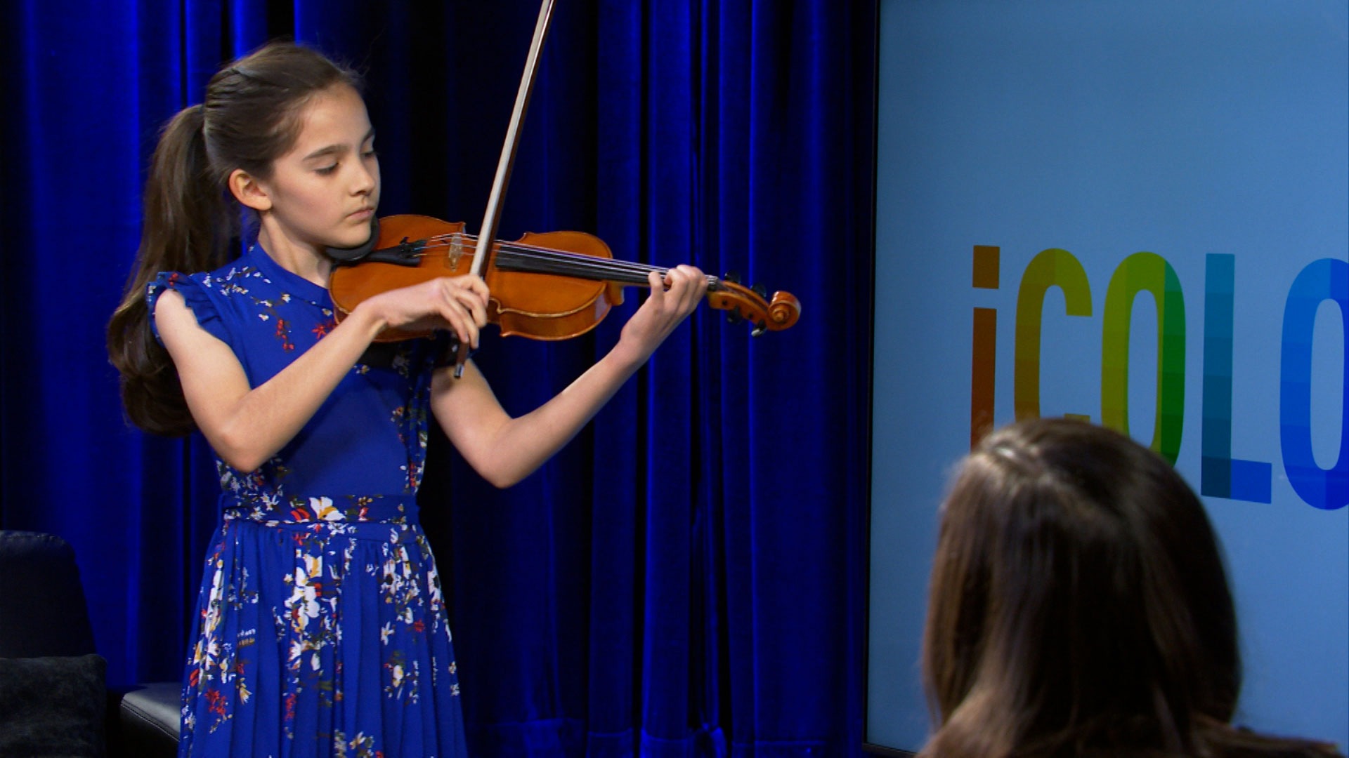 A young girl in a blue floral dress plays the violin on stage, with blue curtains behind her and a colorful logo visible on a screen to the right.