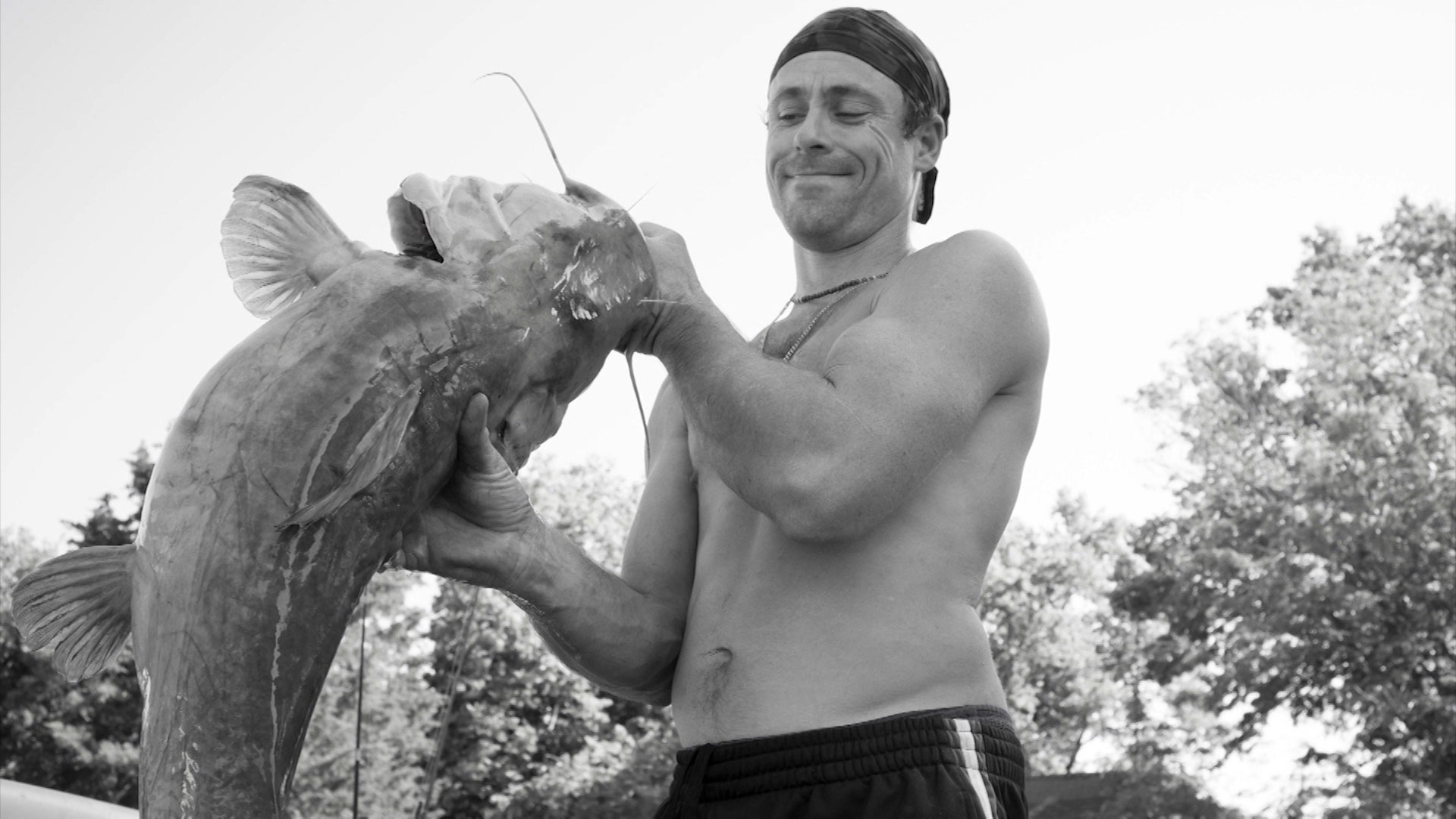 Shirtless man holding a large catfish outdoors with both hands, trees in the background.