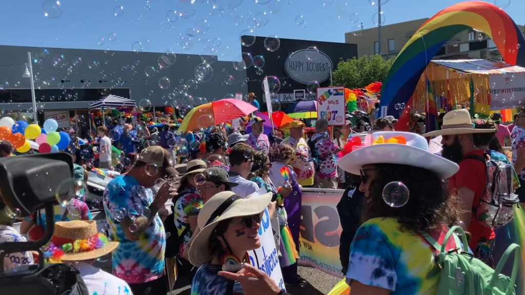A large crowd dressed in tie-dye shirts and hats gathers outside under umbrellas and rainbow decorations, with bubbles floating in the air at a daytime outdoor event.