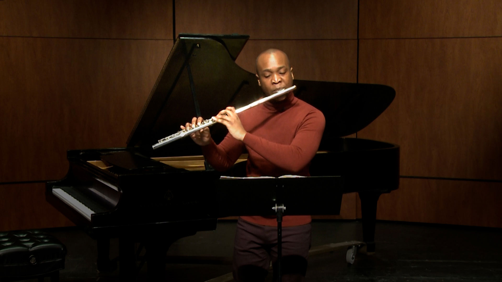 A person in a red long-sleeve shirt plays the flute in front of a grand piano and music stand on a stage with wooden paneling in the background.