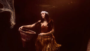 A woman underwater holds a woven basket, wearing a feather headband and a fringed skirt, with fishing nets visible in the background.