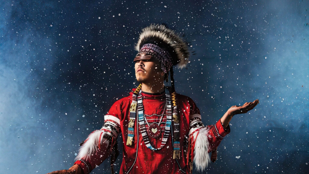 A person in traditional Native American regalia stands with arms raised, looking upward, surrounded by falling snow against a dark blue background.