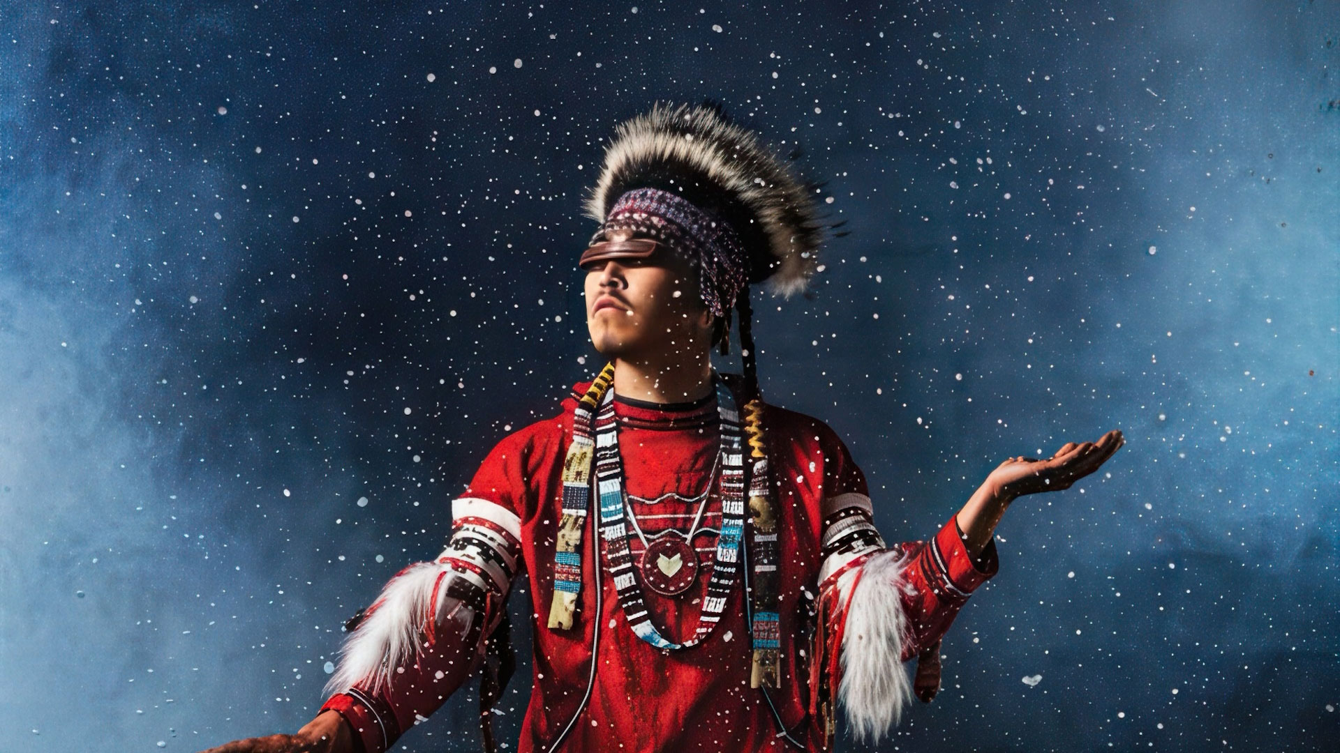 A person in traditional Native American regalia stands with arms raised, looking upward, surrounded by falling snow against a dark blue background.