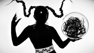 A figure in a glossy black mask with twisted horn-like hair holds a transparent sphere filled with tangled wires, set against a white background.