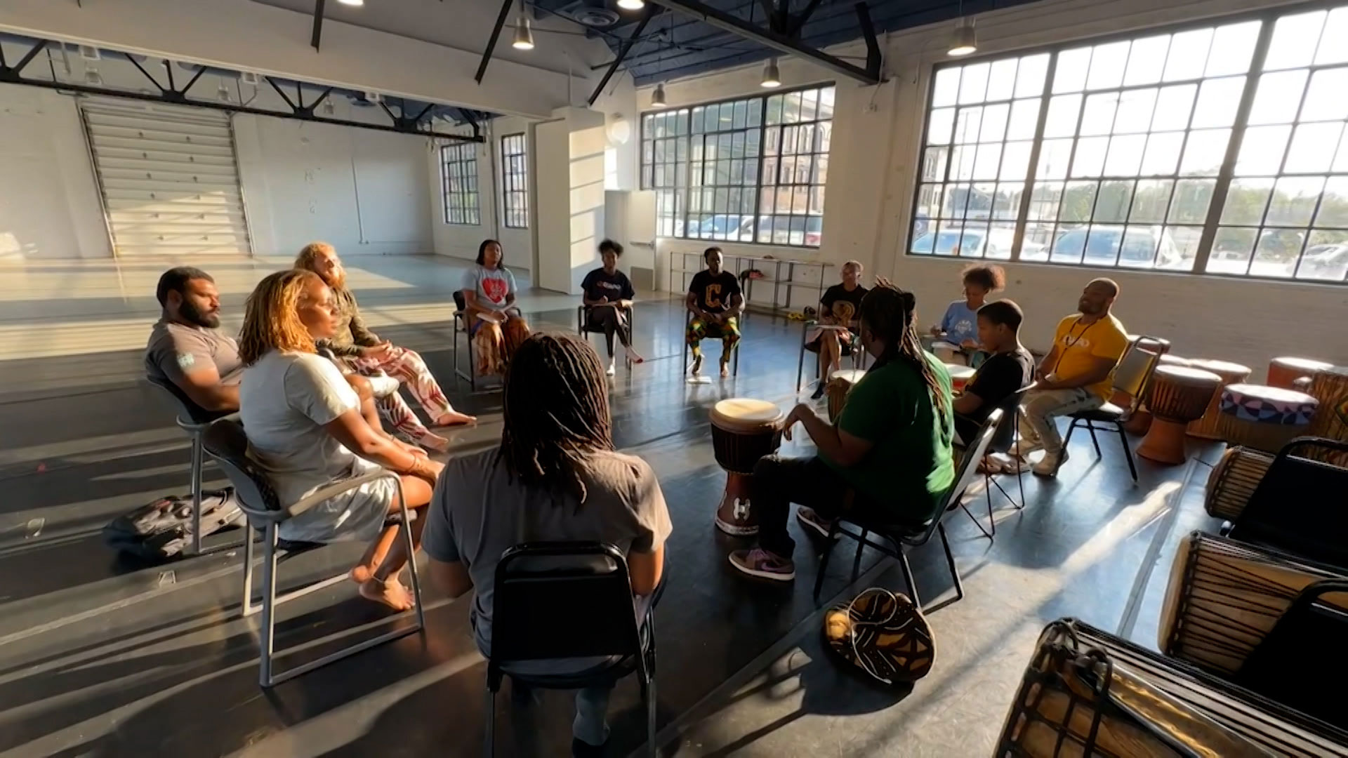 A group of people sit in a circle on chairs in a sunlit room, playing drums and percussion instruments together.