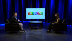 Two people sit in armchairs facing each other on a TV studio set with a screen displaying the word "¡Colores!" in colorful letters.