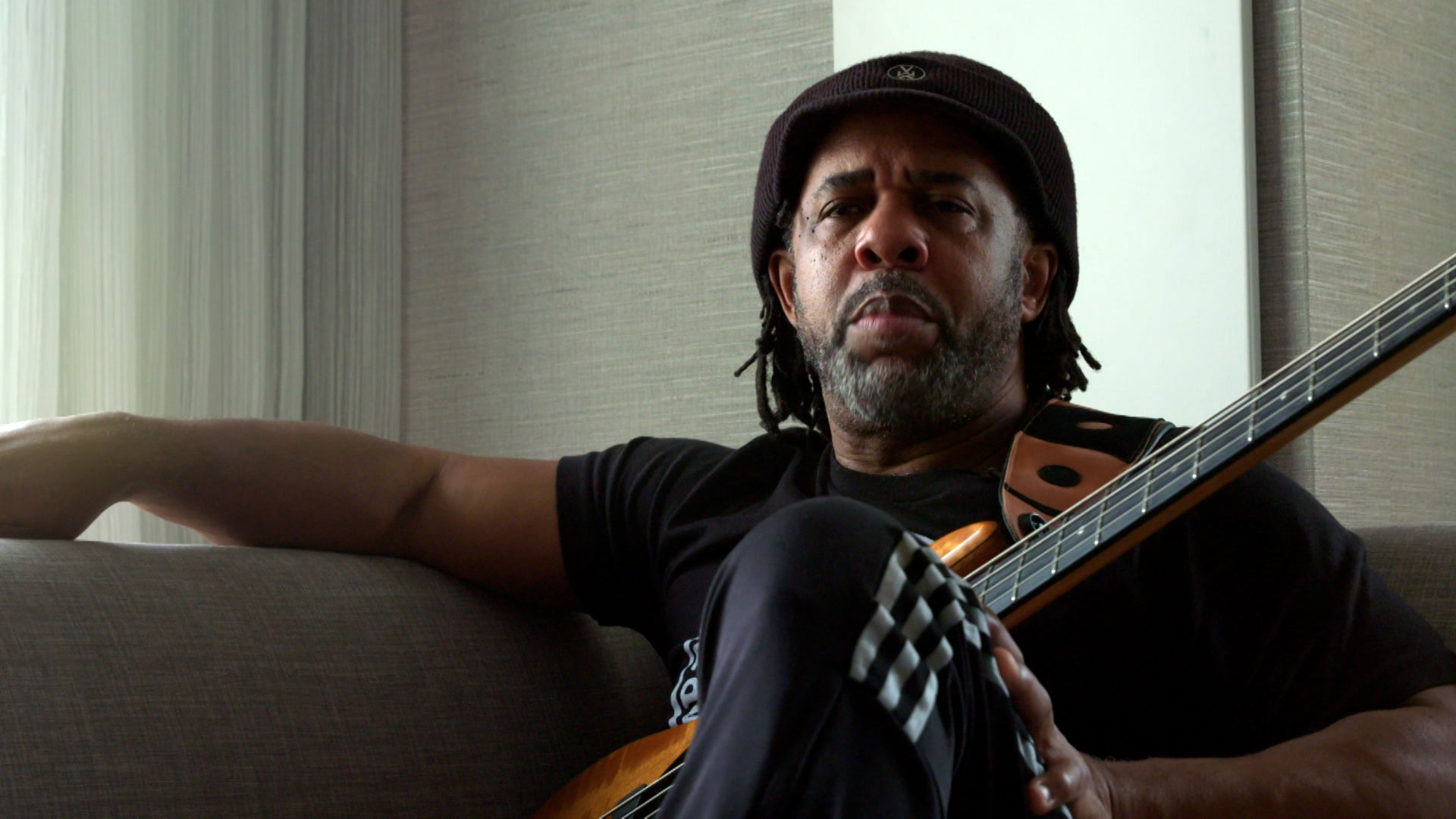 A man with a short beard and hat sits on a couch, holding a bass guitar, looking directly at the camera in a relaxed indoor setting.