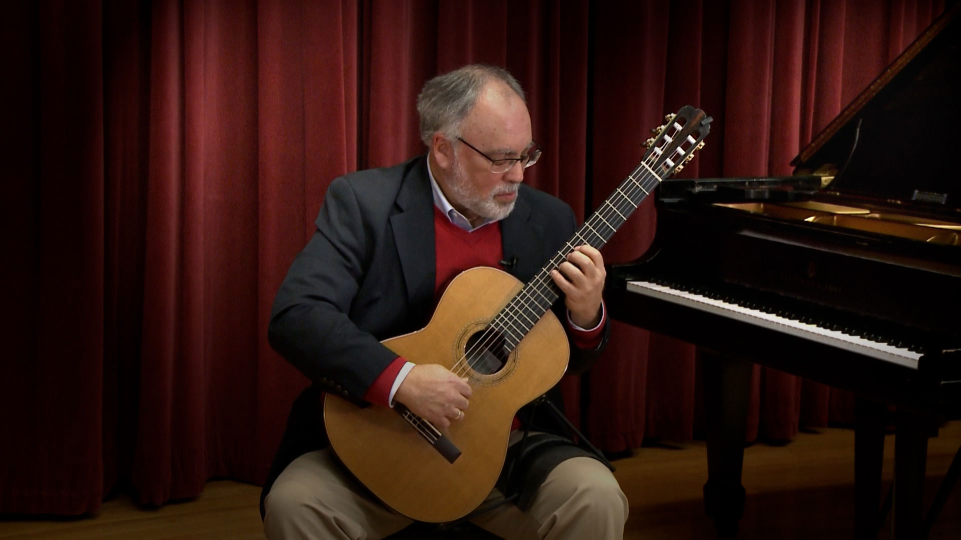 A man with gray hair and a beard plays a classical guitar while seated next to a grand piano in front of red curtains.