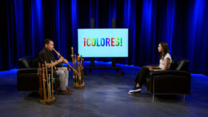 A musician plays wooden flutes while seated across from a woman in a studio with a screen displaying the word “¡COLORES!”.