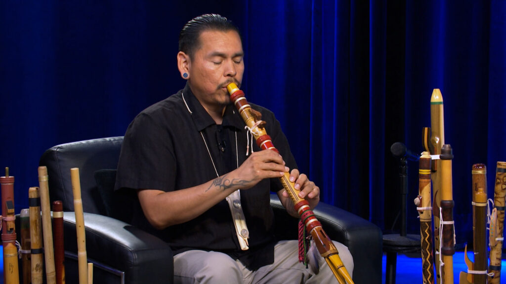 A person sits on a black chair playing a wooden flute, surrounded by several other flutes, with a dark blue curtain in the background.
