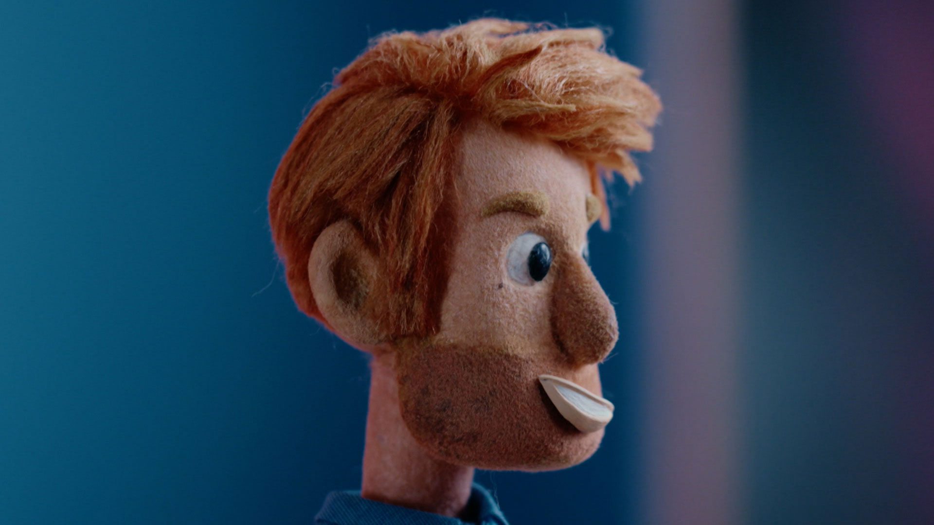 A close-up of a puppet with orange hair, blue eyes, and a slight smile, shown against a blurred blue background.