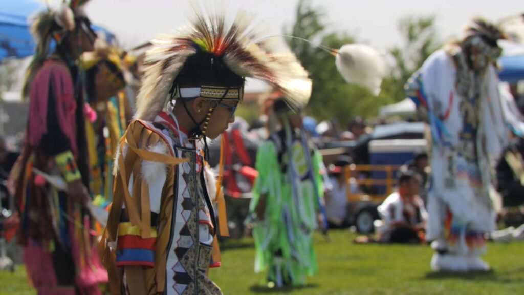 A person in traditional regalia participates in a Native American powwow, with other dancers and attendees visible in the background on a grassy field.
