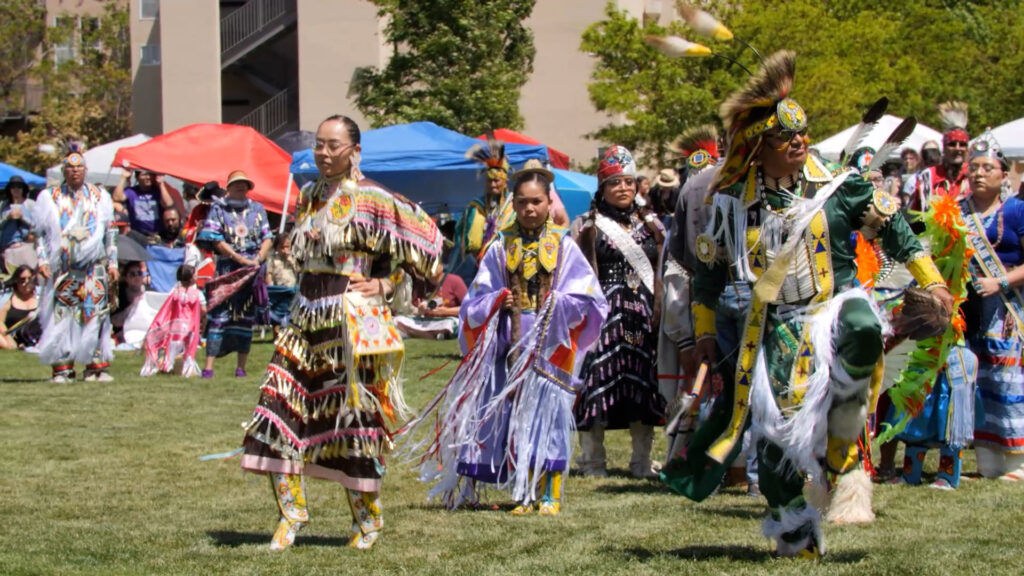 A group of people in colorful traditional Native American regalia dance outdoors on a grassy field during a cultural event.