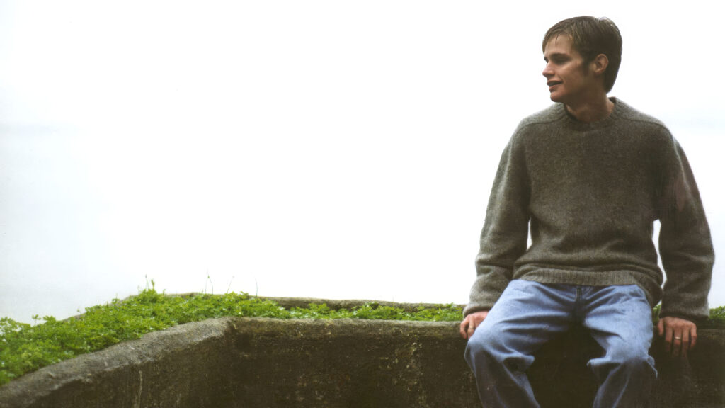 A person wearing a gray sweater and blue jeans sits on the edge of a concrete structure with green plants, looking to the right. The background is bright and mostly white.