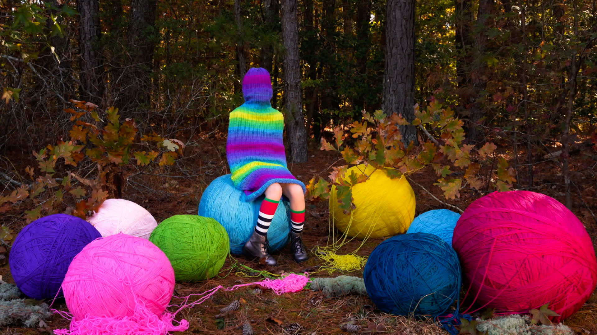 A person covered in a rainbow knit hooded outfit sits on a large blue yarn ball, surrounded by oversized colorful yarn balls in a forest clearing.