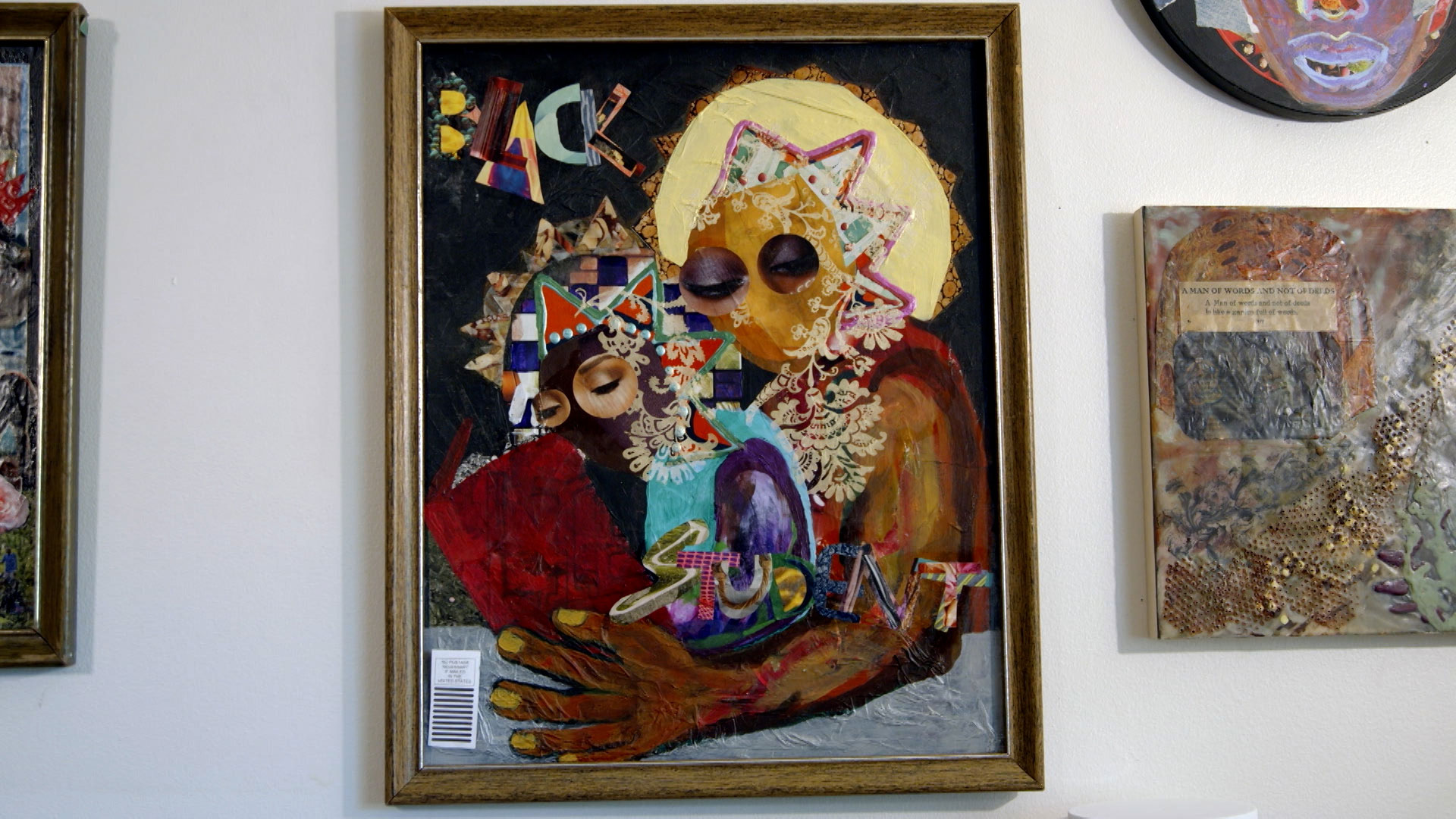 A framed mixed media artwork depicts two figures with intertwined, decorative patterns and the words "BLACK STUDNT" across the piece, hanging on a white wall among other artworks.
