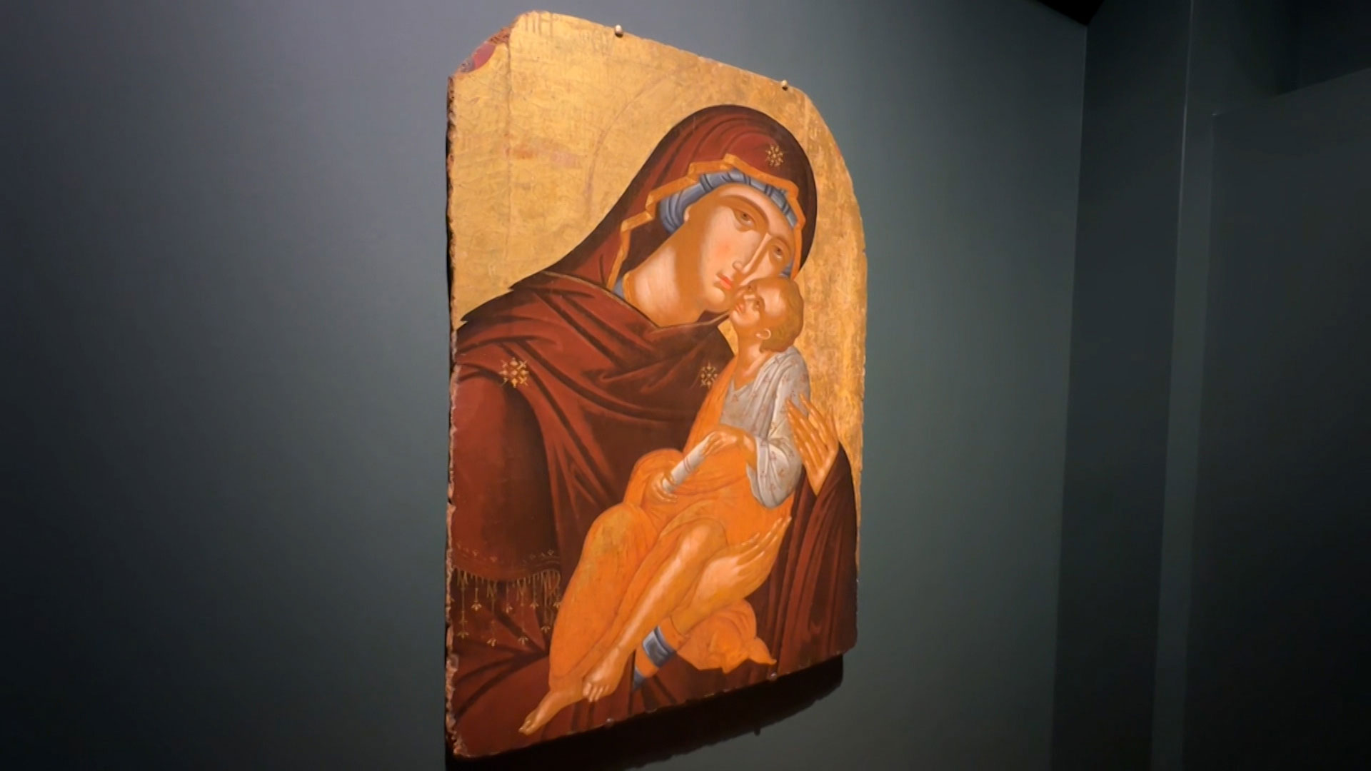 A painted religious icon depicts a woman in a red robe holding a child against a gold background, displayed on a dark blue wall.