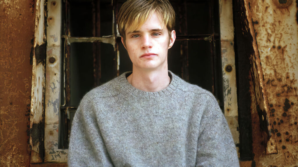 A young person with short blond hair wearing a gray sweater sits in front of a weathered, rusted window frame.
