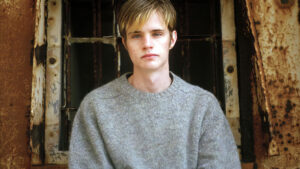 A young person with short blond hair wearing a gray sweater sits in front of a weathered, rusted window frame.