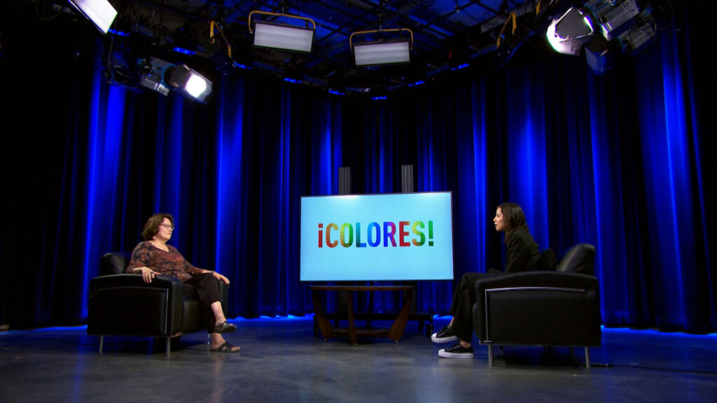 Two people sit in armchairs on a studio set with blue lighting, facing each other with a screen between them displaying the text “¡COLORES!”.