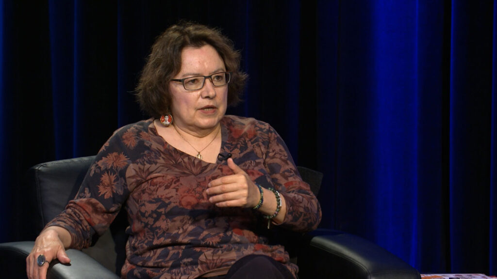 A woman with glasses and shoulder-length hair sits in a chair, gesturing with her hand while speaking against a dark blue curtain background.